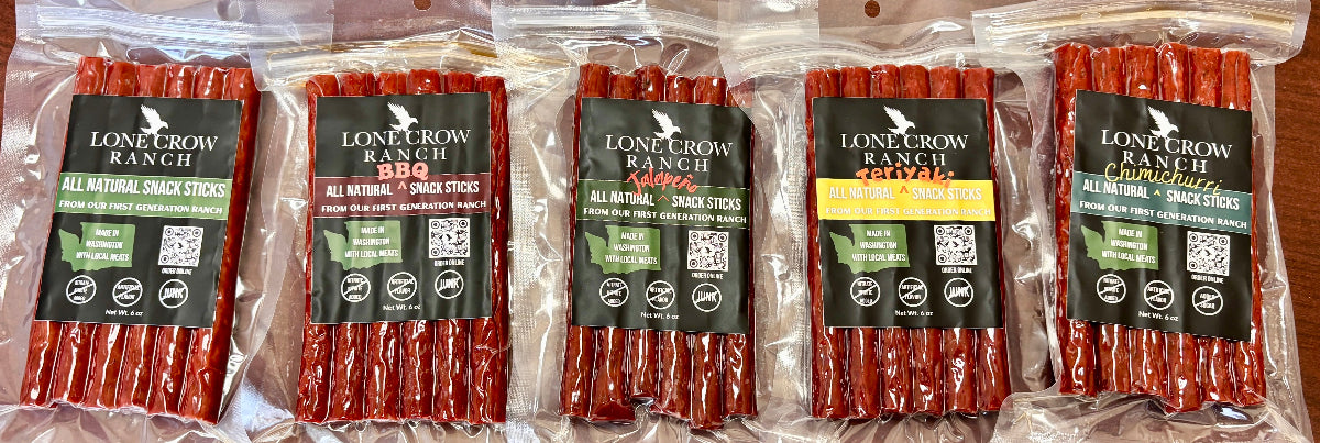 Five packages of Lone Crow Ranch all natural snack sticks arranged side by side on a wooden surface. The packages are clear plastic bags containing reddish-brown snack sticks, with black labels featuring the brand name and product details.