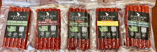 Five packages of Lone Crow Ranch all natural snack sticks arranged side by side on a wooden surface. The packages are clear plastic bags containing reddish-brown snack sticks, with black labels featuring the brand name and product details.