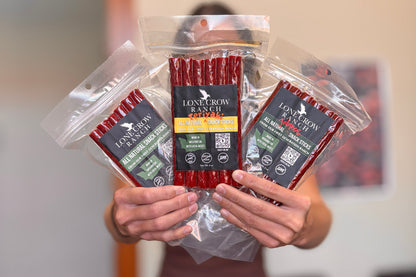 A person holds three packages of Lone Crow Ranch beef jerky sticks in front of their face. The packages are clear plastic with black and yellow labels, showing the red beef jerky sticks inside.
